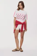 Remera blanca de manga corta, con cuello redondo, calce relajado y estampado frontal con motivos gráficos de soles y símbolos marinos en tonos rosados y bordo, además de líneas horizontales.
