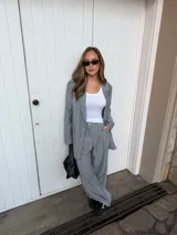 Conjunto de dos piezas compuesto por un blazer y un pantalón de corte sastrero, confeccionados en tela de algodón con estampado de rayas verticales blancas sobre fondo gris. El blazer presenta solapas clásicas y cierre frontal, mientras que el pantalón es de tiro medio y pierna recta.