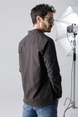 Camisa verde militar de corte recto, con cuello clásico, cierre frontal con botones a presión, mangas largas con puños abotonados, dos bolsillos de parche en el pecho y bajo redondeado.