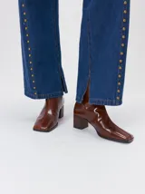 Botas de caña media color marrón, confeccionadas en cuero con acabado brillante. Presentan punta cuadrada, taco de madera y un distintivo bordado decorativo en color rojo que recorre el empeine hasta la punta.