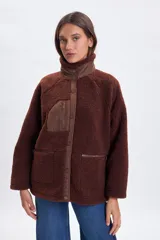 Campera de sherpa color marrón con cuello alto, cierre frontal con botones a presión y detalles en tela técnica a tono. Presenta un bolsillo vertical con cierre en el pecho y dos bolsillos laterales con cierre.