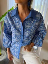 Camisa de manga larga con diseño de flores bordadas en color blanco sobre base azul. Presenta cuello clásico, cierre frontal con botones y mangas abullonadas con puños elásticos.