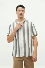 Camisa de manga corta con cuello cubano, confeccionada en una mezcla de lino y algodón con textura. Presenta un diseño a rayas verticales en tonos beige y celeste sobre fondo blanco, con cierre frontal de botones y logo bordado en el pecho.