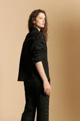 Sobrecamisa negra de estilo estructurado con cierre frontal de botones metálicos, cuello clásico y detalle de tachas decorativas en la zona de los hombros y el pecho. Presenta mangas largas con puños abotonados.