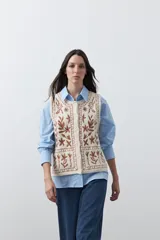 Chaleco tejido color marrón con bordados artesanales de motivos florales y geométricos en color crema. Presenta cuello redondo, cierre frontal con botones y diseño sin mangas.
