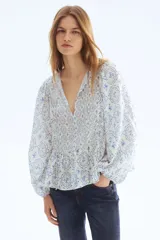 Blusa en mezcla de viscosa con peplum acampanado, cuello redondo, escote en V y tiras finas con cuentas decorativas. Presenta mangas globo largas con elástico en los puños y un estampado en tonos blancos y azules con motivos florales y geométricos.