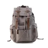 Mochila de tela canvas color gris con detalles en símil cuero marrón, tapa con correas de hebilla y cierre superior tipo marinero. Incluye múltiples bolsillos exteriores con cierre, compartimento interno para laptop de 15 pulgadas y correas ajustables.