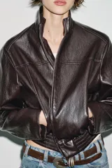 Chaqueta estilo bomber corta de efecto piel en color marrón oscuro, con cuello subido, cierre frontal con cremallera y cintura elástica fruncida.