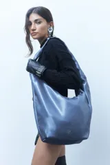 Bolso tipo hobo de cuero vegano con acabado brillante metalizado en tono azulado. Presenta una estructura blanda, asa corta integrada y un pequeño aplique metálico con logo en la parte inferior.