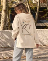 Canguro de felpa color crudo, con capucha, corte amplio, bolsillos delanteros y aberturas laterales en el dobladillo. Presenta un estampado en la espalda con diseño de arco y flecha rodeado de estrellas, y detalles gráficos en las mangas.