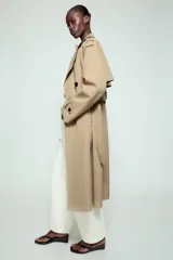 Trench largo de color negro con cuello de solapa, manga larga con trabillas ajustables y cierre frontal cruzado con doble botonadura. Cuenta con bolsillos delanteros de vivo, solapa cortavientos en el pecho y cinturón ajustable del mismo tejido.