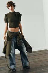 Jersey de punto de manga corta y cuello redondo, en color verde oliva. Presenta un detalle de lazo grande en el frente que le otorga un toque distintivo y femenino.