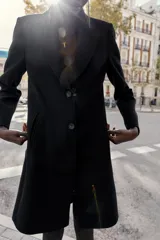 Abrigo de paño color beige con cuello solapa, manga larga con hombreras y cierre frontal con botones. Presenta bolsillos delanteros de doble vivo y abertura en la espalda.