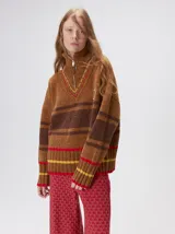 Pullover de punto con textura aterciopelada, diseño oversize, cuello polo con cierre frontal parcial y detalles de pespuntes en contraste. Presenta rayas horizontales en tonos tierra y un parche bordado con la letra M en el pecho.