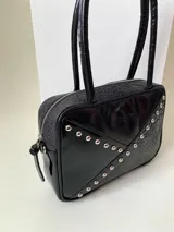 Cartera rígida negra con detalles en rojo y tachas plateadas. Tiene herrajes y cierre metálico plateado marca YKK, asa fina para llevar al hombro o en la mano y correa larga extra regulable. El interior es forrado, tiene un bolsillo con cierre enmarcado en cuero, y un segundo bolsillo plaqué.