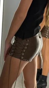 Short de cuero marrón oscuro con efecto desgastado, tiro bajo, cierre frontal con botón plateado, detalles de cordones ajustables en los laterales y tachas plateadas en el dobladillo.