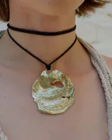 Collar artesanal con dije central de metal con textura orgánica irregular que evoca una ostra. El dije está montado sobre un cordón de algodón natural, permitiendo un uso versátil y regulable.