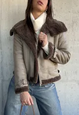 Campera tipo gamulán de cuero sintético en color vizón, con interior y solapas de piel sintética en color marrón oscuro. Presenta cierre frontal, correas ajustables con hebillas en los puños y diseño de corte recto.