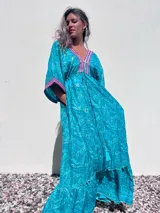 Túnica larga de estilo boho, color turquesa con estampado paisley en tonos azules y dorados. Presenta escote en V profundo con bordados decorativos en color fucsia y blanco, mangas amplias tipo kimono y ajuste en la cintura con cordón. El diseño incluye un ruedo con volado y terminaciones con borlas.