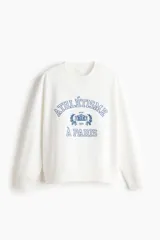 Buzo oversize color blanco de cuello redondo y mangas largas, con interior cepillado suave. Presenta un estampado frontal en tono azul con texto de estilo universitario y diseño decorativo. Cuenta con ribetes acanalados en el cuello, los puños y el bajo.