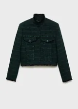 Chaqueta de tweed en color verde oscuro, con diseño corto y recto. Presenta cuello alto, cierre frontal con botones, mangas largas y dos bolsillos de solapa con botón en el frente. Se caracteriza por sus terminaciones deshilachadas y costuras decorativas.