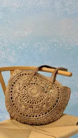 Bolso playero grande de tejido de papel trenzado color paja, con forma redonda y asas de cuero marrón. Presenta un forro interior y un dije colgante con motivos marinos.