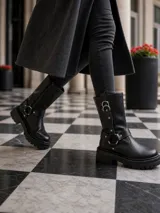 Botas de caña media en cuero negro, con suela track dentada y taco cuadrado. Presentan un diseño con correas ajustables, argollas metálicas y detalles de tachas plateadas.