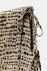 Falda pantalón de tiro bajo con estampado de puntos negros sobre fondo beige. Presenta un diseño de superposición con cierre lateral mediante lazadas y cremallera oculta, finalizando en bordes con detalle de franja negra.