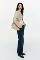 Campera acolchada de nylon beige con diseño guateado, cuello redondo y cierre frontal. Cuenta con hombros caídos, mangas largas, bolsillos laterales y cordón de ajuste en la cintura con tiras colgantes.