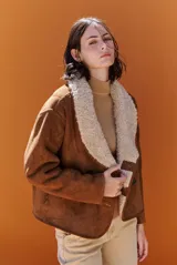 Chaqueta tipo gamulán de gamuza marrón con interior y solapas de corderito sintético en color crema. Presenta un diseño de corte recto con cierre frontal de botones y bolsillos laterales.