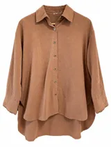 Camisa oversize color marrón chocolate, de manga larga con puños anchos decorados con pequeños pompones, cuello camisero clásico y cierre frontal de botones. Presenta diseño de corte asimétrico, más larga en la parte trasera.