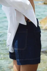 Shorts de denim azul oscuro, tiro alto, con cierre de botón dorado. Los bolsillos delanteros tienen un detalle de ribete trenzado.