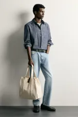 Bolso tipo shopper de tejido color crudo, con doble asa de hombro y detalles en cuero negro. Cuenta con compartimento principal con cierre de tira y botón a presión.