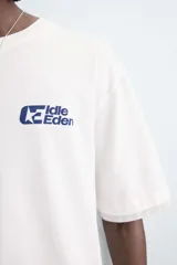 Camiseta de manga corta y cuello redondo, color blanco roto, con efecto de doble capa (la capa inferior gris asoma en el ruedo). Presenta estampados pequeños en azul marino en el pecho: un logo con texto 'Idle Eden' y otros símbolos.