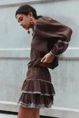 Blusa de algodón con textura arrugada, color marrón, con detalles bordados en contraste en el frente y las mangas. Presenta escote redondo, cierre frontal con botones, mangas globo largas y puños abotonados.