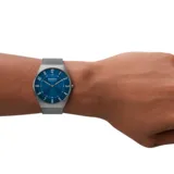Reloj analógico de cuarzo con caja de acero inoxidable en tono gris carbón y malla metálica tipo milanesa. Presenta una esfera azul con acabado sunray, números arábigos y dos agujas.