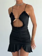 Vestido corto negro de tirantes finos, con escote pronunciado y abertura central decorada con volados de encaje. El cuerpo es ajustado y presenta frunces laterales que terminan en un ruedo con volado.