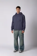 Buzo canguro oversize de frisa de algodón premium en color azul grisáceo. Presenta un diseño con recortes frontales, bolsillo tipo canguro y un pequeño logo circular estampado en el pecho. Cuenta con puños y ruedo acanalados.