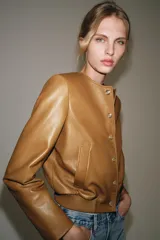 Campera bomber de cuero 100% color tostado de la ZARA WOMAN COLLECTION, con cuello redondo, manga larga, bolsillos delanteros ribeteados y cierre frontal con botones metálicos en relieve. Presenta elástico fruncido en el dobladillo inferior.