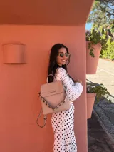 Mochila tipo cartera con tapa, confeccionada en símil cuero color beige. Presenta un diseño moderno con detalle de ojales metálicos en la parte frontal, compartimentos internos organizadores y bolsillo trasero con cierre.