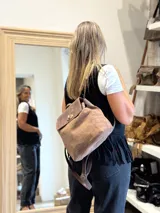 Mochila de cuero color marrón con solapa superior en tono marrón oscuro. Presenta un cierre metálico giratorio y detalles de cordones cruzados en la parte inferior frontal.