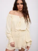Vestido corto color crema, de diseño off-shoulder con mangas largas abullonadas, cintura ajustada con frunces elásticos y falda corta de volados.