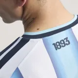 Camiseta de fútbol Adidas de la Selección Argentina, modelo titular 2026, versión jugador. Diseño a rayas verticales celestes y blancas, con cuello redondo y detalles en azul marino en las mangas y hombros. Incluye escudo de la AFA y logo de Adidas en dorado, además del parche de campeón FIFA.