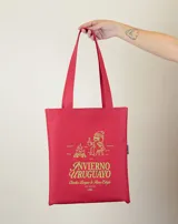 Tote bag de tela color rojo con estampado gráfico en color verde lima que representa una fogata y una persona sentada tomando una bebida caliente, acompañado por el texto 'Invierno Uruguayo'.