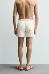 Short de baño en tejido técnico color beige claro, con cintura elástica ajustable con cordón y botón, bolsillos laterales y detalle de costuras en contraste.