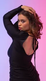 Vestido midi de color negro, confeccionado en tejido con acabado satinado. Presenta un diseño ajustado al cuerpo con cuello bote, mangas largas y detalle de frunces laterales a lo largo de la falda.