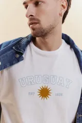 Remera de manga corta color blanco con cuello redondo. Presenta un estampado frontal con la palabra 'URUGUAY' en tipografía universitaria, un sol central en color dorado y la inscripción 'EST 1825'.