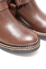 Bota de caña alta de cuero marrón, con diseño urbano y líneas limpias. Cuenta con una correa decorativa en el tobillo con argolla metálica y remaches plateados, suela de goma con costura reforzada y tirador trasero superior.