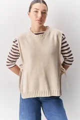 Chaleco tejido de punto color beige, con cuello redondo, terminaciones en rib y lazos laterales regulables que permiten ajustar la prenda.