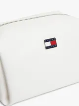 Cartera bandolera blanca Tommy Jeans de poliuretano, diseño cuadrado con cierre de cremallera y compartimento principal. Cuenta con correa ajustable, insignia metálica con el logo de la marca en el frente y un colgante lateral extraíble de cordón rojo trenzado con argollas metálicas.